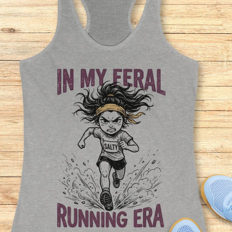 Gray tank top with graphic design of a running woman and text Mint green tank top with graphic design and text "in my feral running era" on a wooden surface
