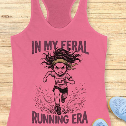 Pink tank top with graphic design of a running woman and text  Mint green tank top with graphic design and text "in my feral running era" on a wooden surface 