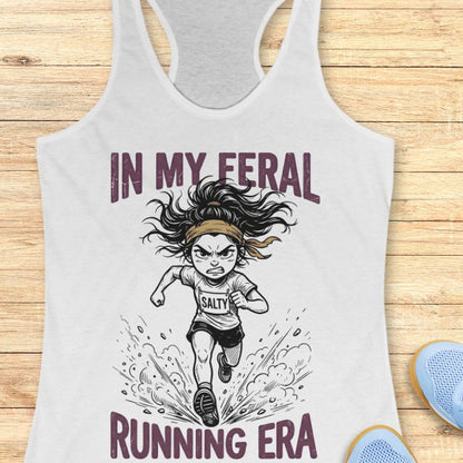 White tank top with graphic design and text Mint green tank top with graphic design and text "in my feral running era" on a wooden surface
