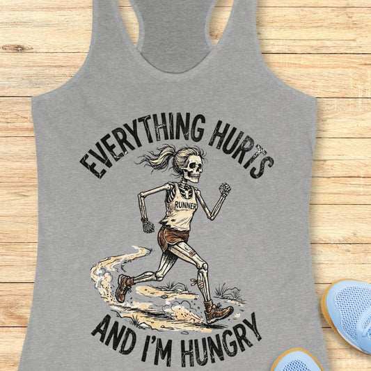 Gray tank top with skeleton runner graphic and text 'Everything hurts and I'm hungry' on a wooden surface.