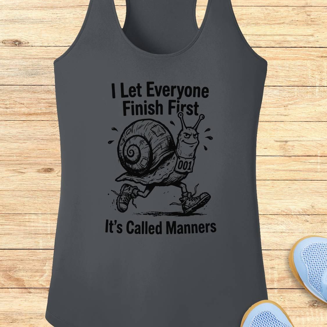 A dark gray performance racerback tank top laid flat on a light wood background, featuring a black graphic of a snail wearing a race bib and the text 'I Let Everyone Finish First' and 'It's Called Manners'.