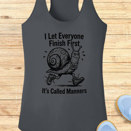 A dark gray performance racerback tank top laid flat on a light wood background, featuring a black graphic of a snail wearing a race bib and the text 'I Let Everyone Finish First' and 'It's Called Manners'.