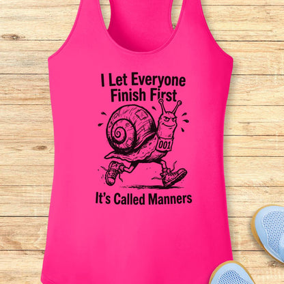 A bright pink racerback tank top laid flat on a light wood surface, featuring a black graphic of a snail wearing shoes and the text 'I Let Everyone Finish First' and 'It's Called Manners.' Partial blue shoes are visible at the bottom right as styling props.