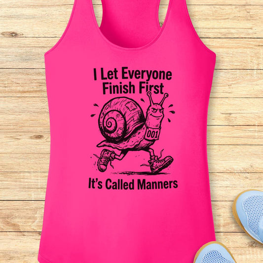 A bright pink racerback tank top laid flat on a light wood surface, featuring a black graphic of a snail wearing shoes and the text 'I Let Everyone Finish First' and 'It's Called Manners.' Partial blue shoes are visible at the bottom right as styling props.
