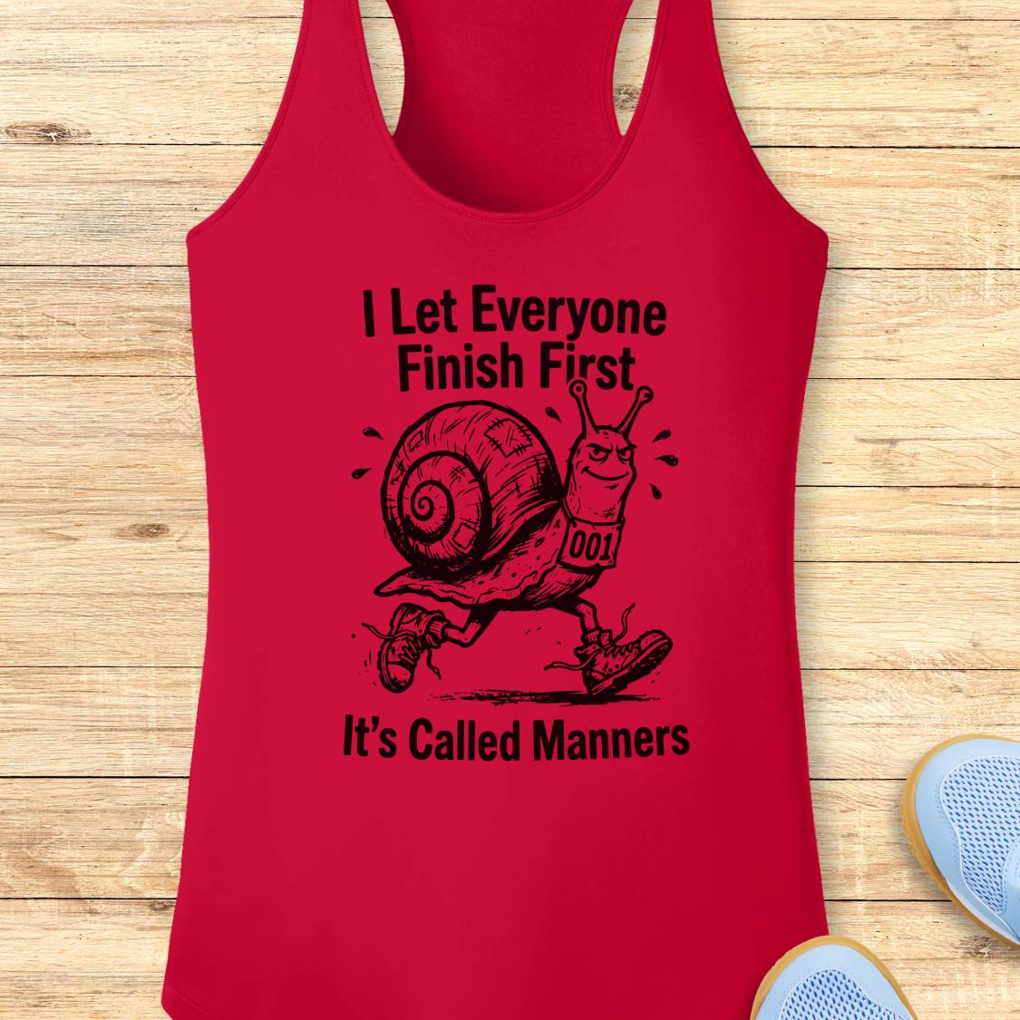 A red performance racerback tank top with a black graphic of a snail running and the text 'I Let Everyone Finish First It's Called Manners', displayed flat on a light wood background with partial shoes visible at the edges.