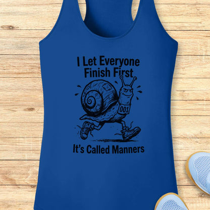 A royal blue performance racerback tank top with a black graphic of a snail running and the text 'I Let Everyone Finish First It's Called Manners', displayed flat on a light wood background with partial sneakers visible at the bottom right.