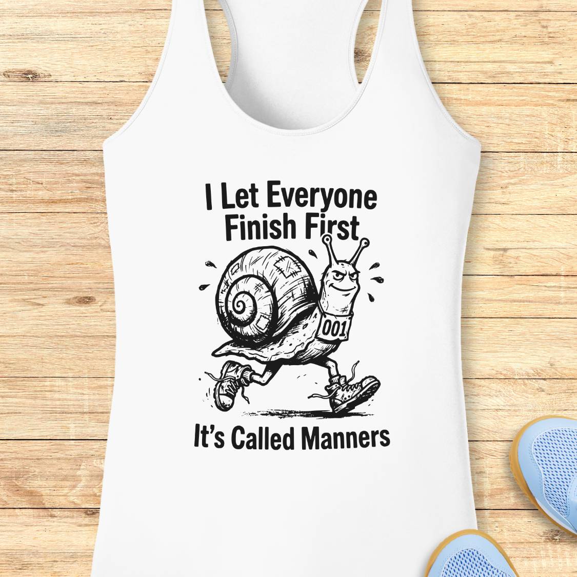 White performance racerback tank top laid flat on a light wood background, featuring a black graphic of a snail wearing a race bib and the text 'I Let Everyone Finish First It's Called Manners.' A partial blue shoe is visible at the bottom right.