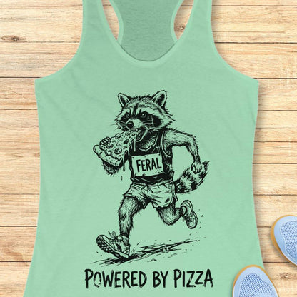 mint tank top with a raccoon runner, with a bib that says - feral, and it is eating a slice of pizza. The tank top has the text  -powered by pizza, and it's on a wooden background