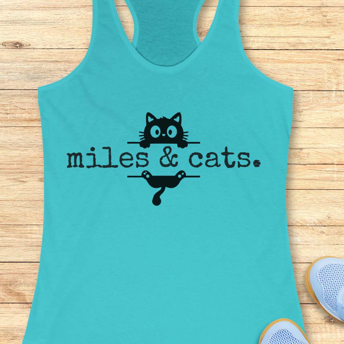 Turquoise tank top with 'miles & cats' text and cat illustrations on a wooden surface.