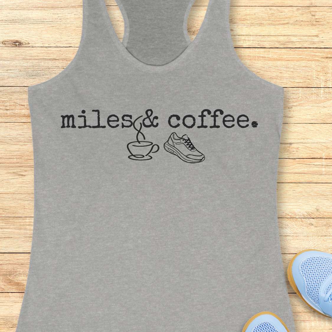 Gray tank top with 'miles & coffee.' text and graphics on a wooden surface.