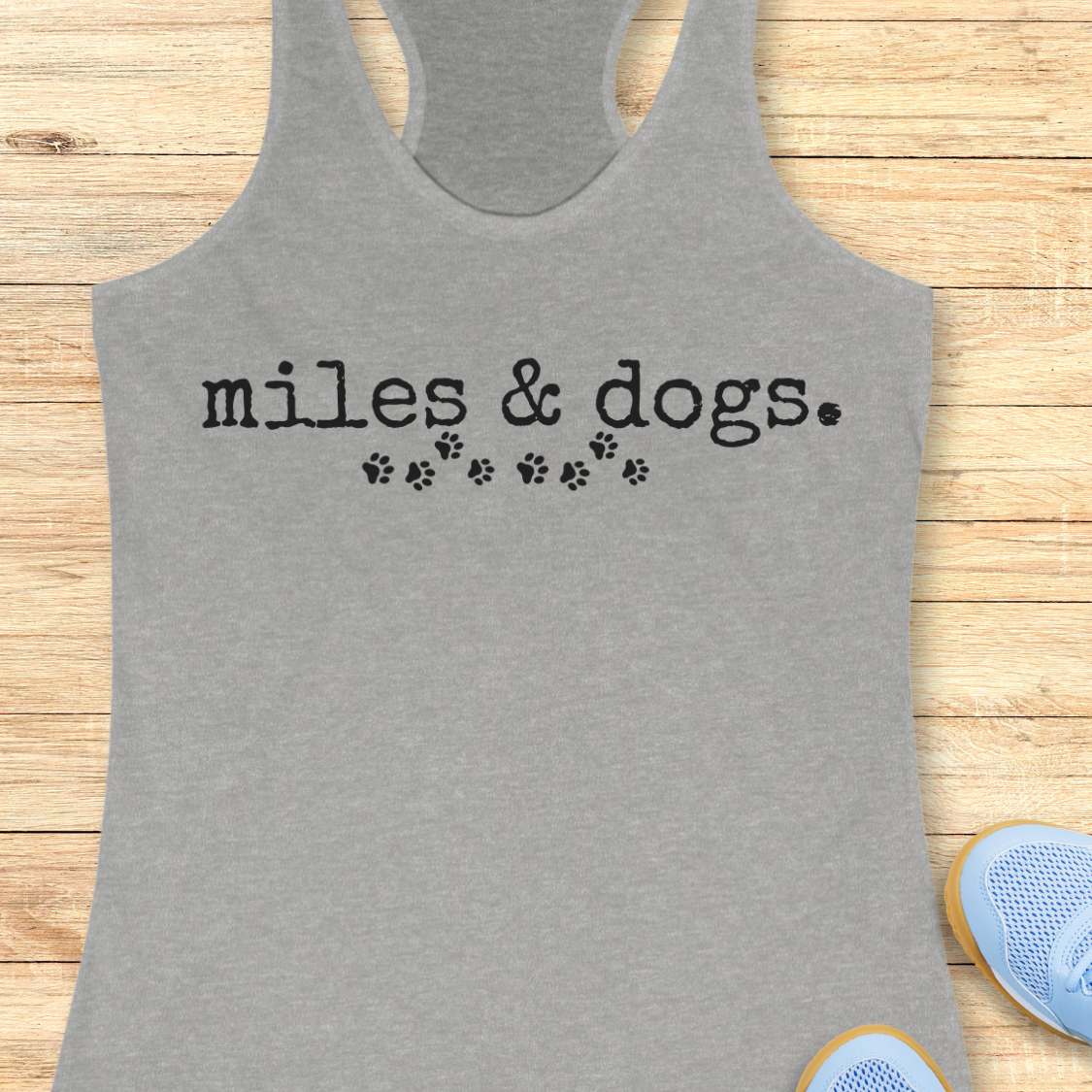 Gray tank top with 'miles & dogs.' text on a wooden surface