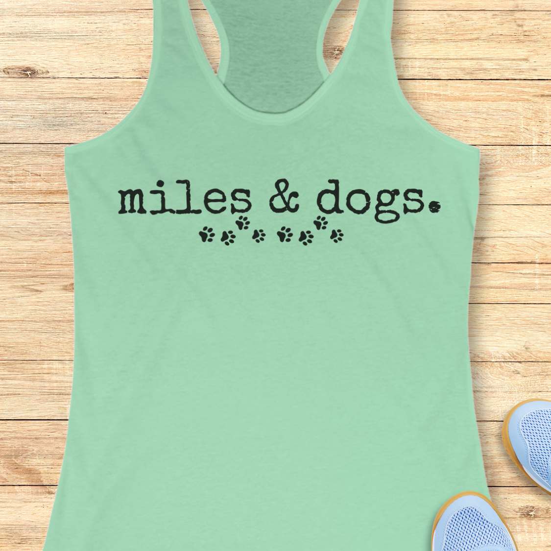 Mint green tank top with 'miles & dogs.' text on a wooden surface