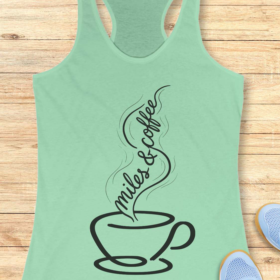 Mint green tank top with a coffee cup design and 'miles & coffee' text on a wooden surface.