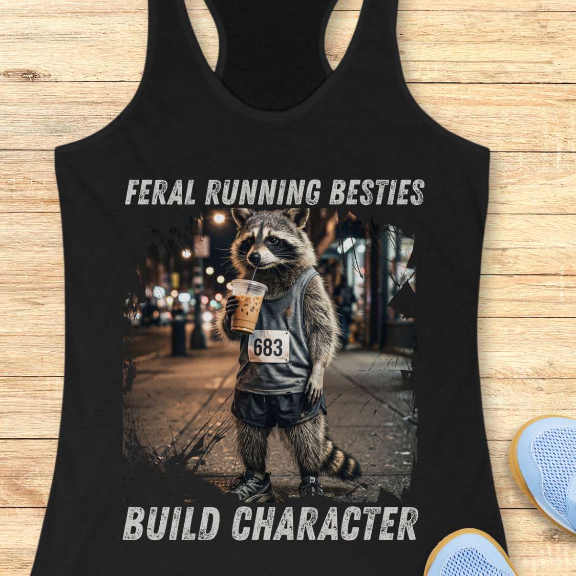 Black tank top with a graphic of a raccoon holding a coffee cup and text  "feral running besties build character" on a wooden surface.