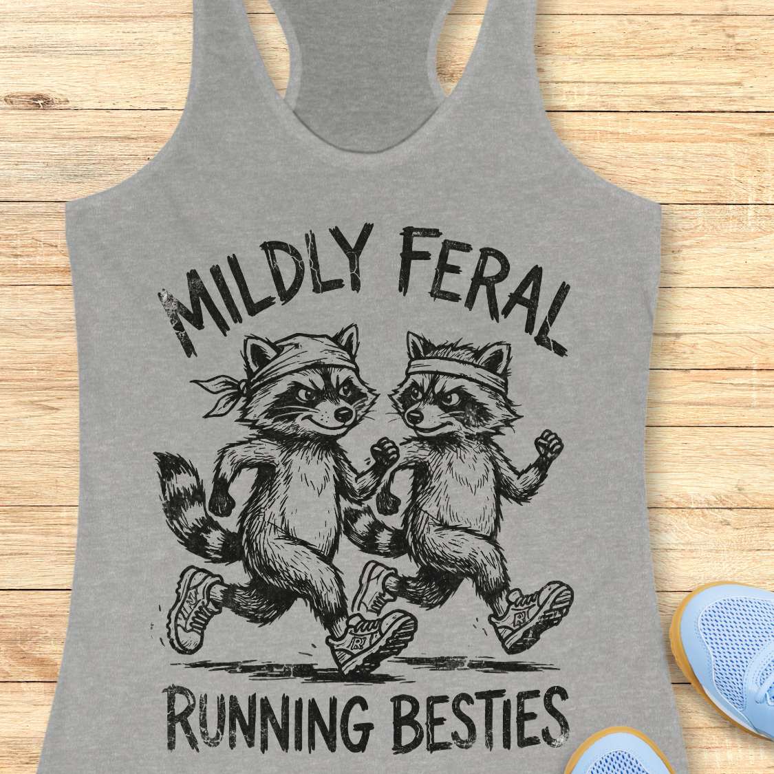 Gray tank top with raccoon design and text 'Mildly Feral Running Besties' on a wooden surface.