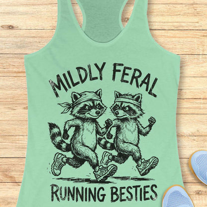 Mint green tank top with raccoon design and text 'Mildly Feral Running Besties' on a wooden surface