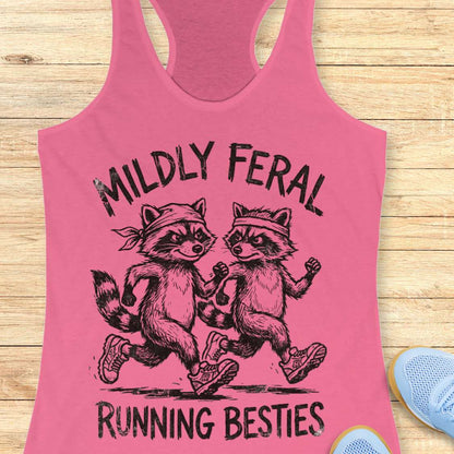 Pink tank top with raccoon design and text 'Mildly Feral Running Besties' on a wooden surface.