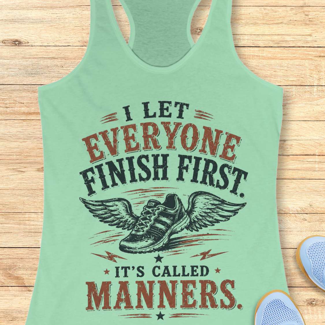 Light green tank top with humorous text "I let everyone finish first. It's called manners." design on a wooden surface