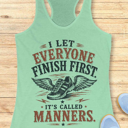 Light green tank top with humorous text "I let everyone finish first. It's called manners." design on a wooden surface