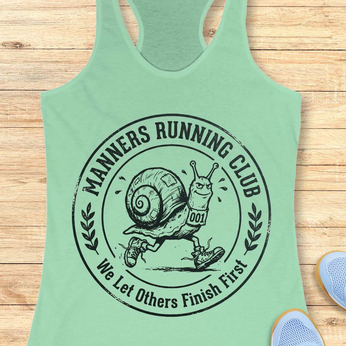 Mint green tank top with a snail graphic and 'Manners Running Club' text on a wooden surface.