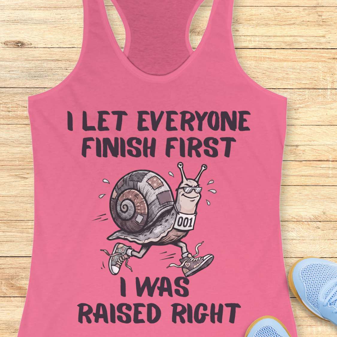 Pink tank top with a snail graphic and text I let everyone finish first I was raised right, on a wooden surface