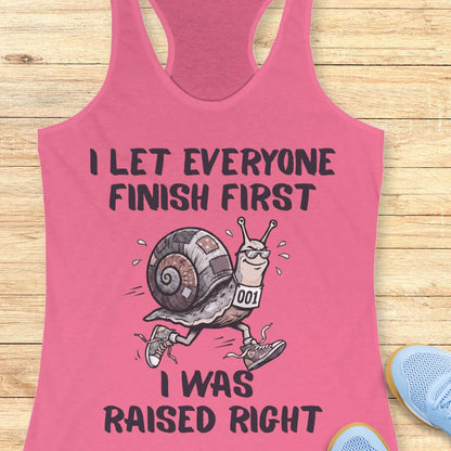 Pink tank top with a snail graphic and text I let everyone finish first I was raised right, on a wooden surface