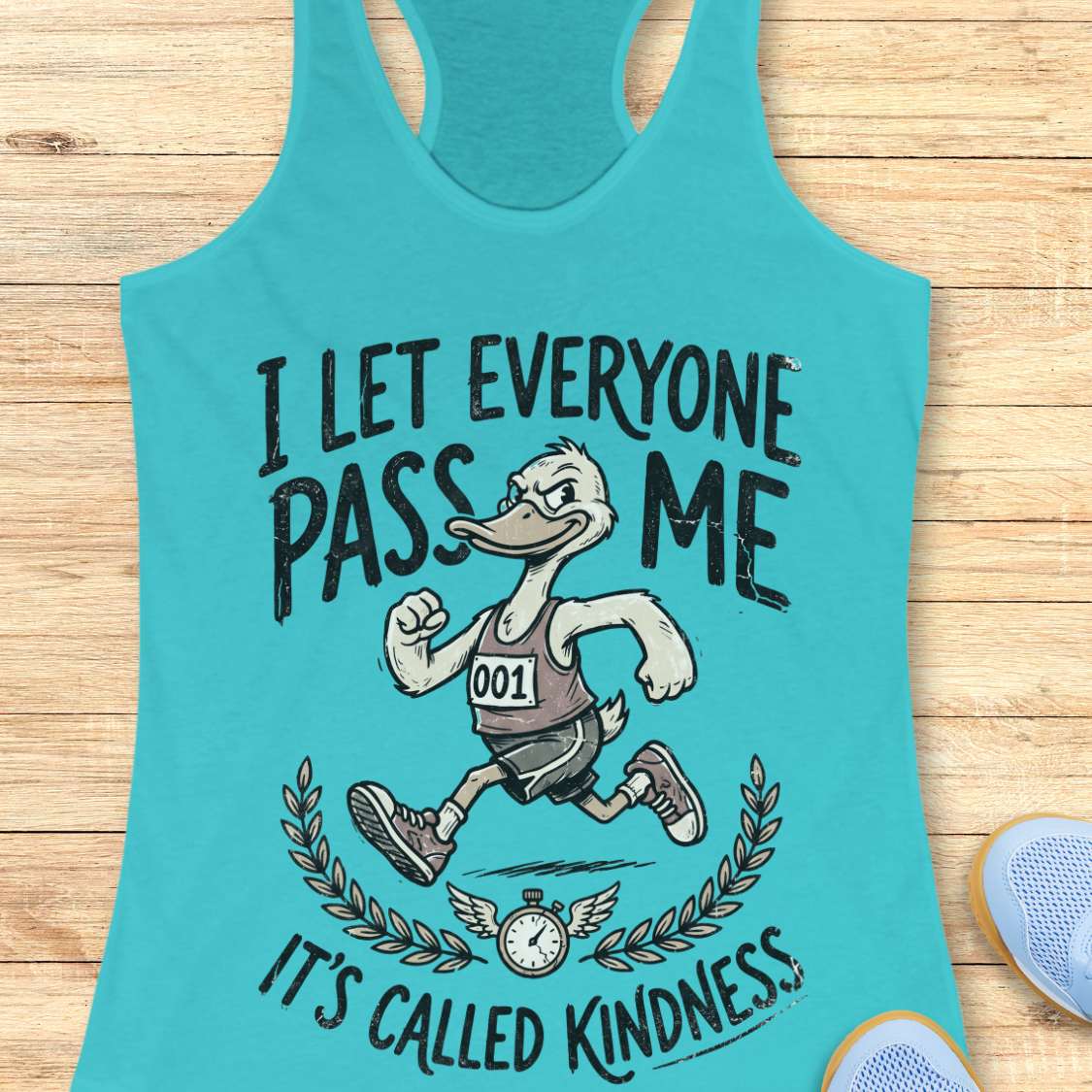 Turquoise tank top with a graphic of a duck running and text I let everyone pass me, it's called kindness, on a wooden surface