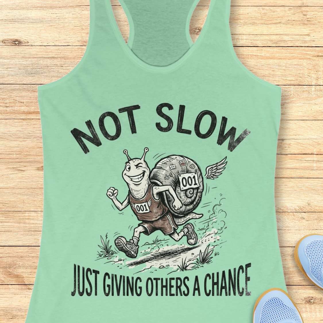 Mint green tank top with a snail graphic and text - not slow, just giving others a chance, on a wooden surface