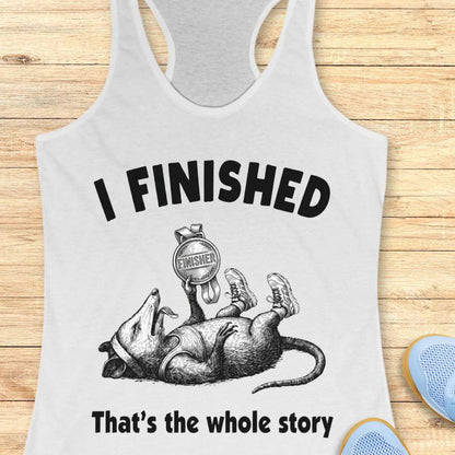 white tank top with graphic of a finished runner and text 'I FINISHED That's the whole story' on a wooden surface.