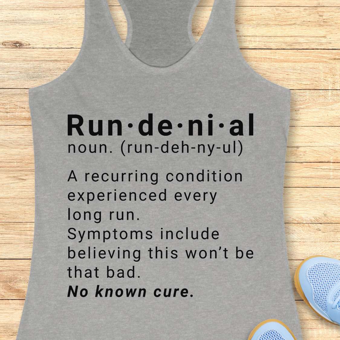 Gray tank top with humorous definition text  of run de ni al  on a wooden surface
