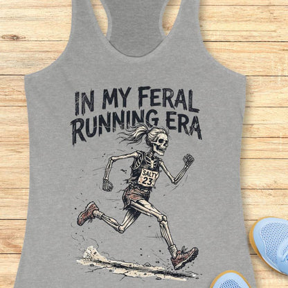 Gray tank top with skeleton running graphic and text 'In my feral running era' on a wooden surface.