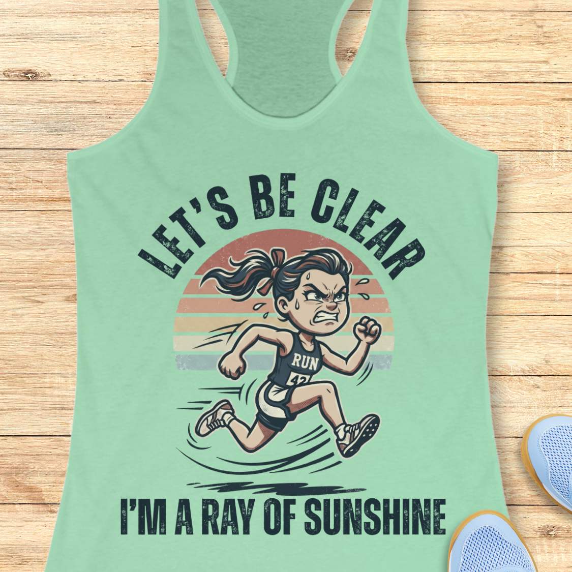 Light green tank top with a graphic of a running person and text let's be clear, I'm a ray of sunshine on a wooden surface