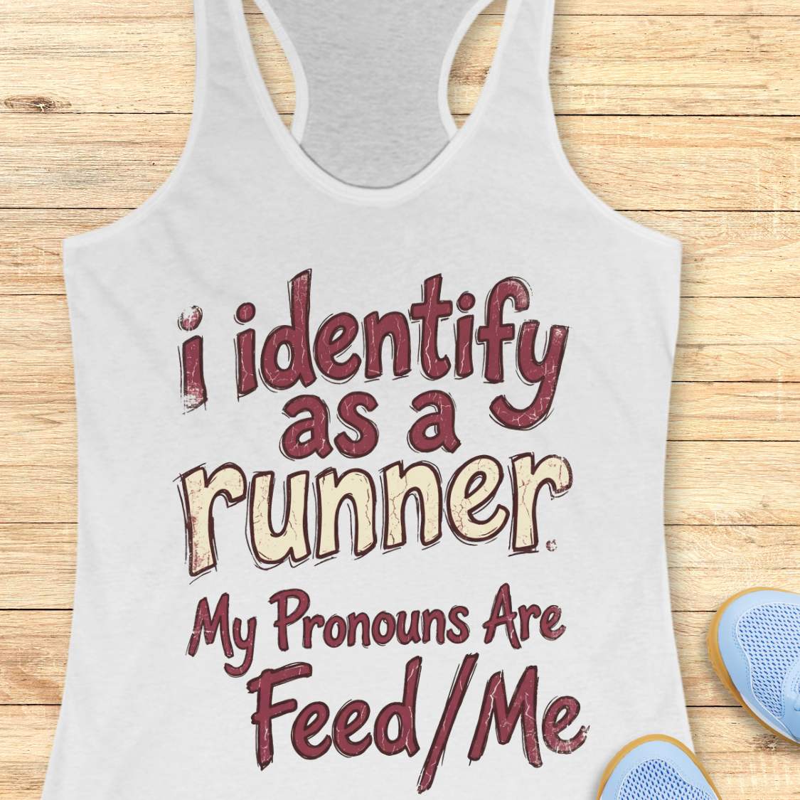 White tank top with text 'i identify as a runner. My pronouns are feed/me' on a wooden surface.