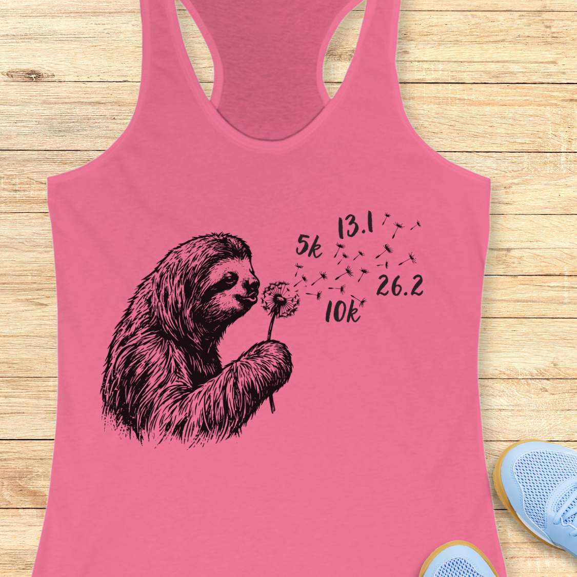 Pink tank top with a sloth blowing a dandelion design on a wooden surface.