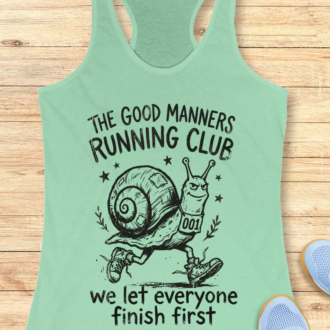 Mint green tank top with a snail graphic and text The good manners running club, we let everyone finish first, on a wooden surface