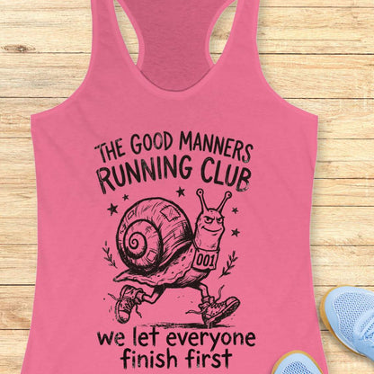 Pink tank top with a snail graphic and text The good manners running club, we let everyone finish first, on a wooden surface