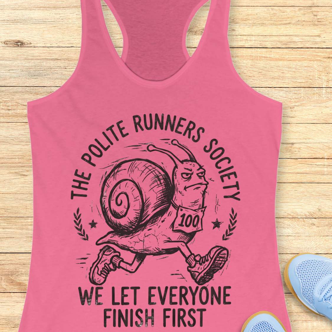 Pink tank top with a snail graphic and text 'The Polite Runners Society' on a wooden surface
