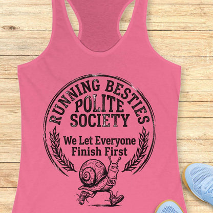 Pink tank top with 'Running Besties Polite Society' text and snail graphic on a wooden surface.