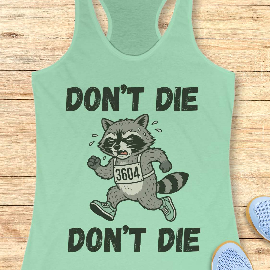 Light green tank top with a raccoon graphic and text 'Don't Die' on a wooden surface