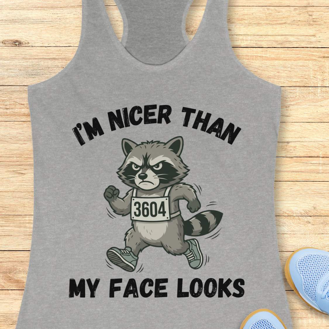 Gray tank top with a raccoon graphic and text I'm nicer than my face looks, on a wooden surface