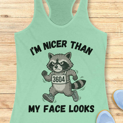 Light green tank top with a raccoon graphic and text I'm nicer than my face looks, on a wooden surface
