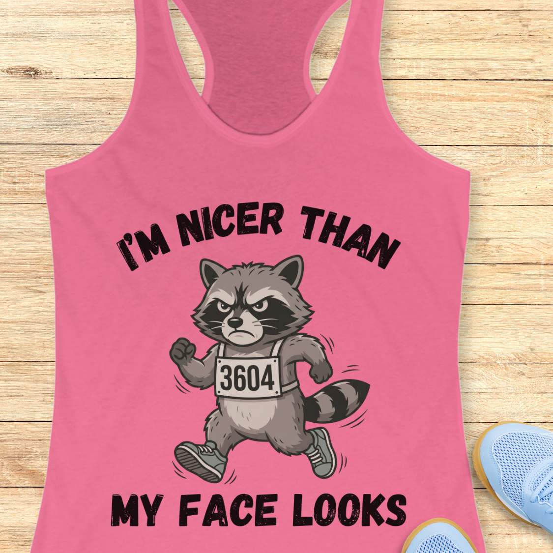 Pink tank top with a raccoon graphic and text I'm nicer than my face looks, on a wooden surface