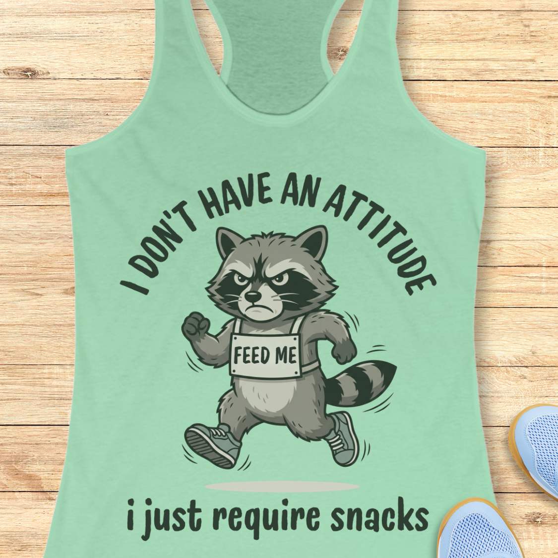 Mint green tank top with a raccoon graphic and text I don't have an attitude, I just require snacks, on a wooden surface