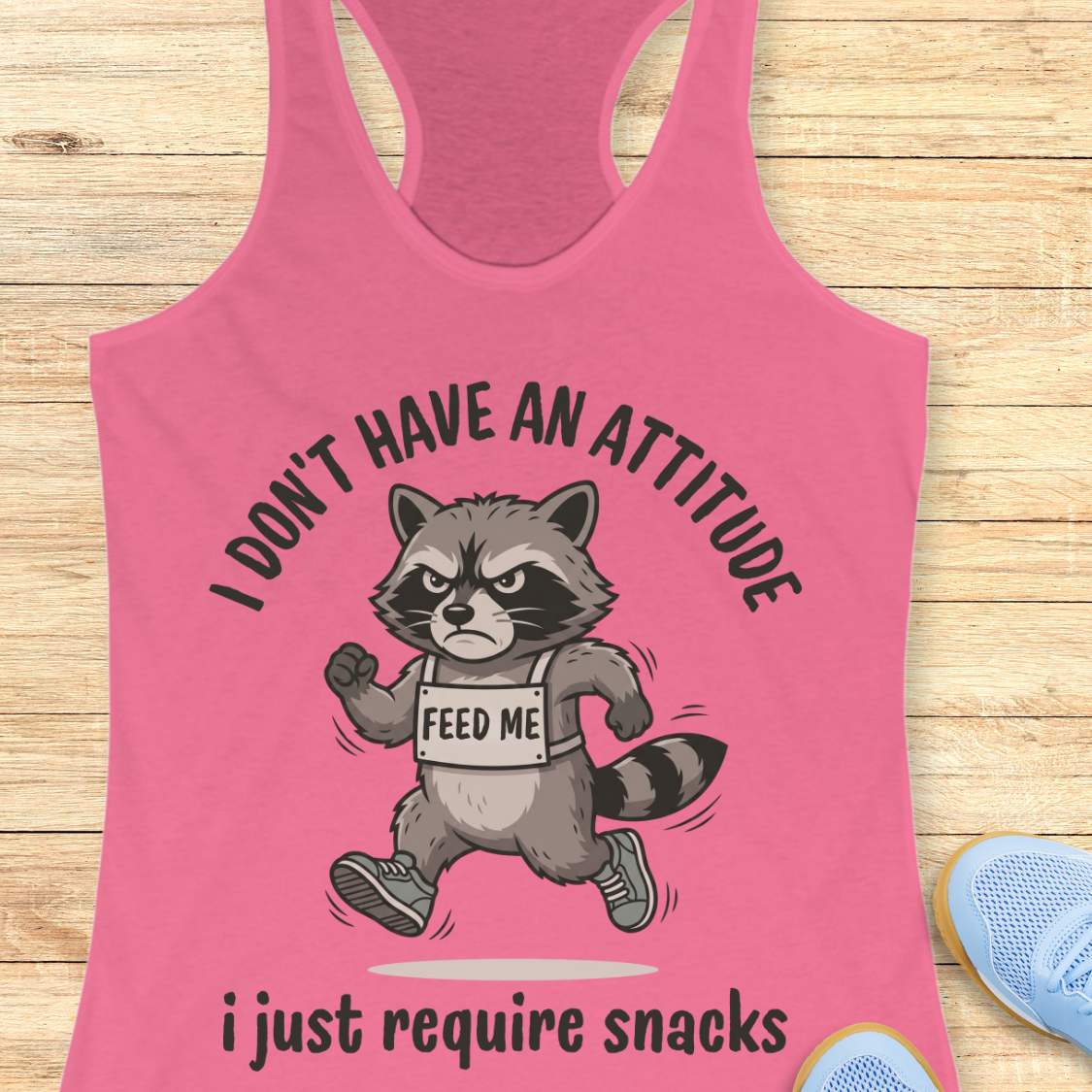 Pink tank top with a raccoon graphic and text I don't have an attitude, I just require snacks, on a wooden surface