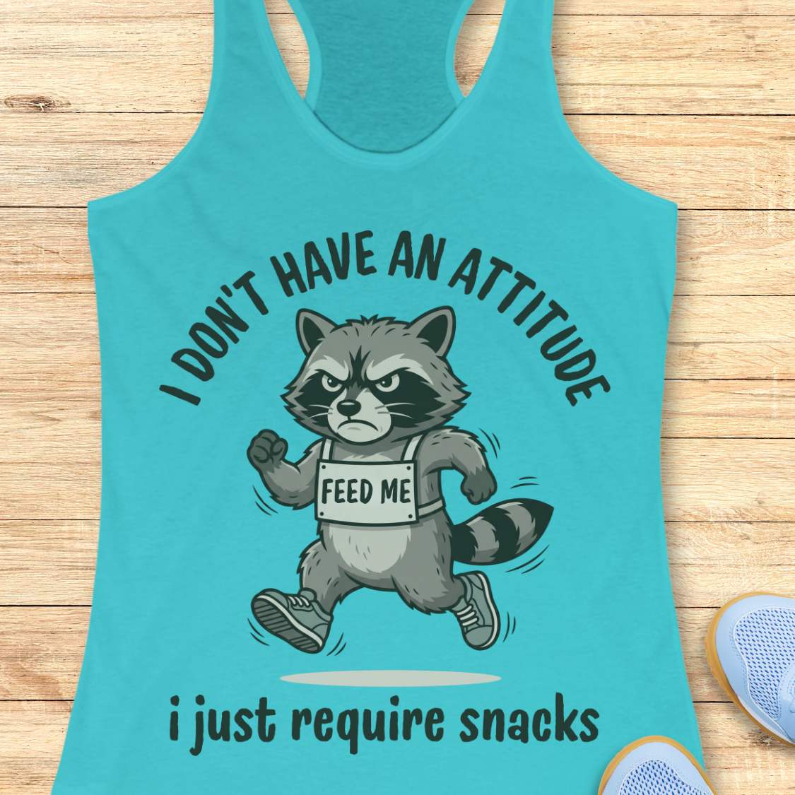 Turquoise tank top with a raccoon graphic and text I don't have an attitude, I just require snacks,  on a wooden surface