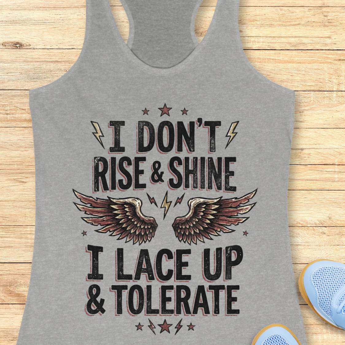 Gray tank top with motivational text I don't rise & shine, I lace up & tolerate,  and wings design on a wooden surface