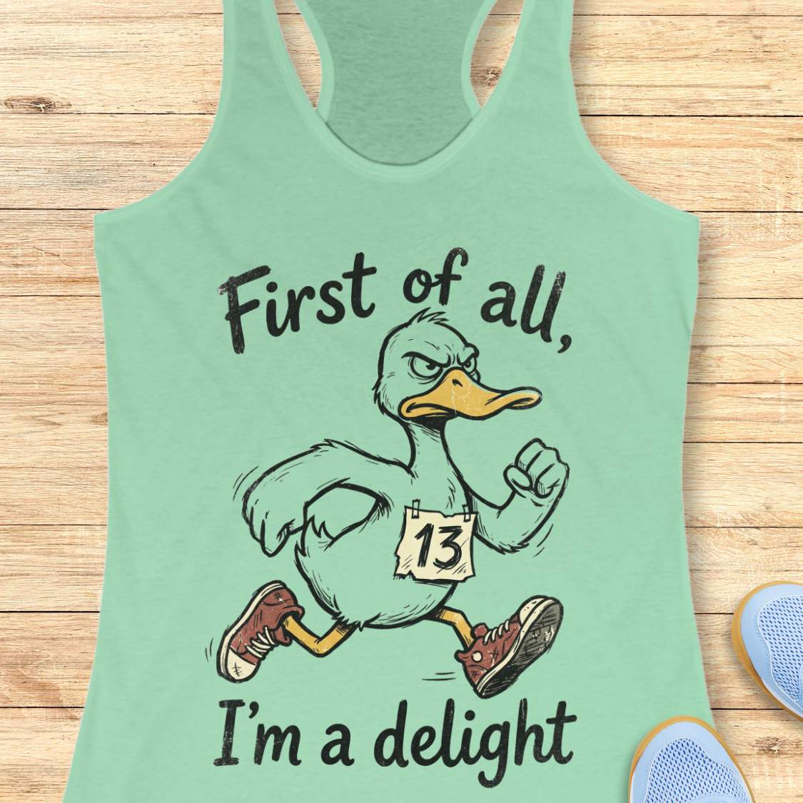 Light green tank top with a cartoon duck and text first of all, I'm a delight, on a wooden surface