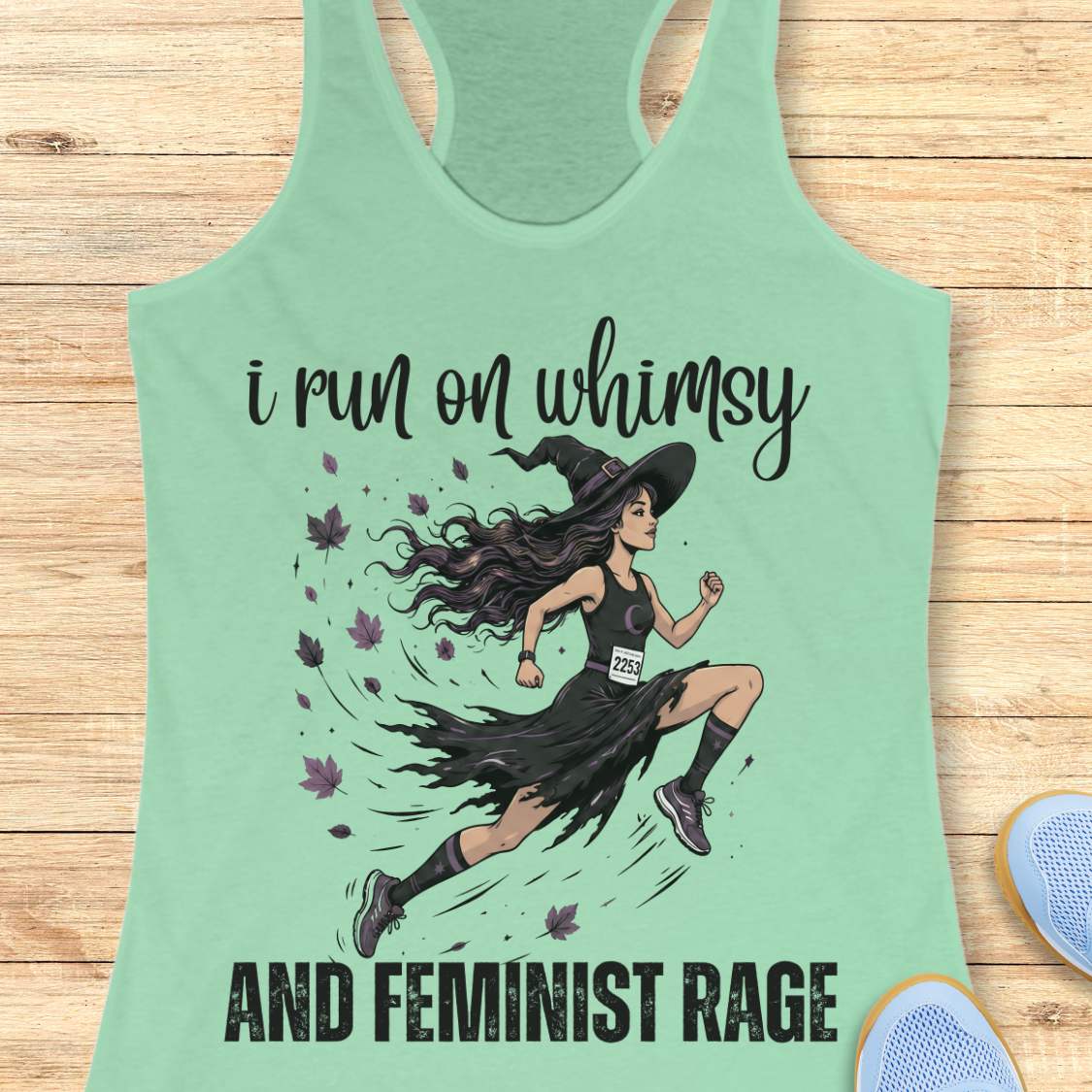 Green tank top with a graphic of a witch runner and text i run on whimsy and feminist rage, on a wooden surface