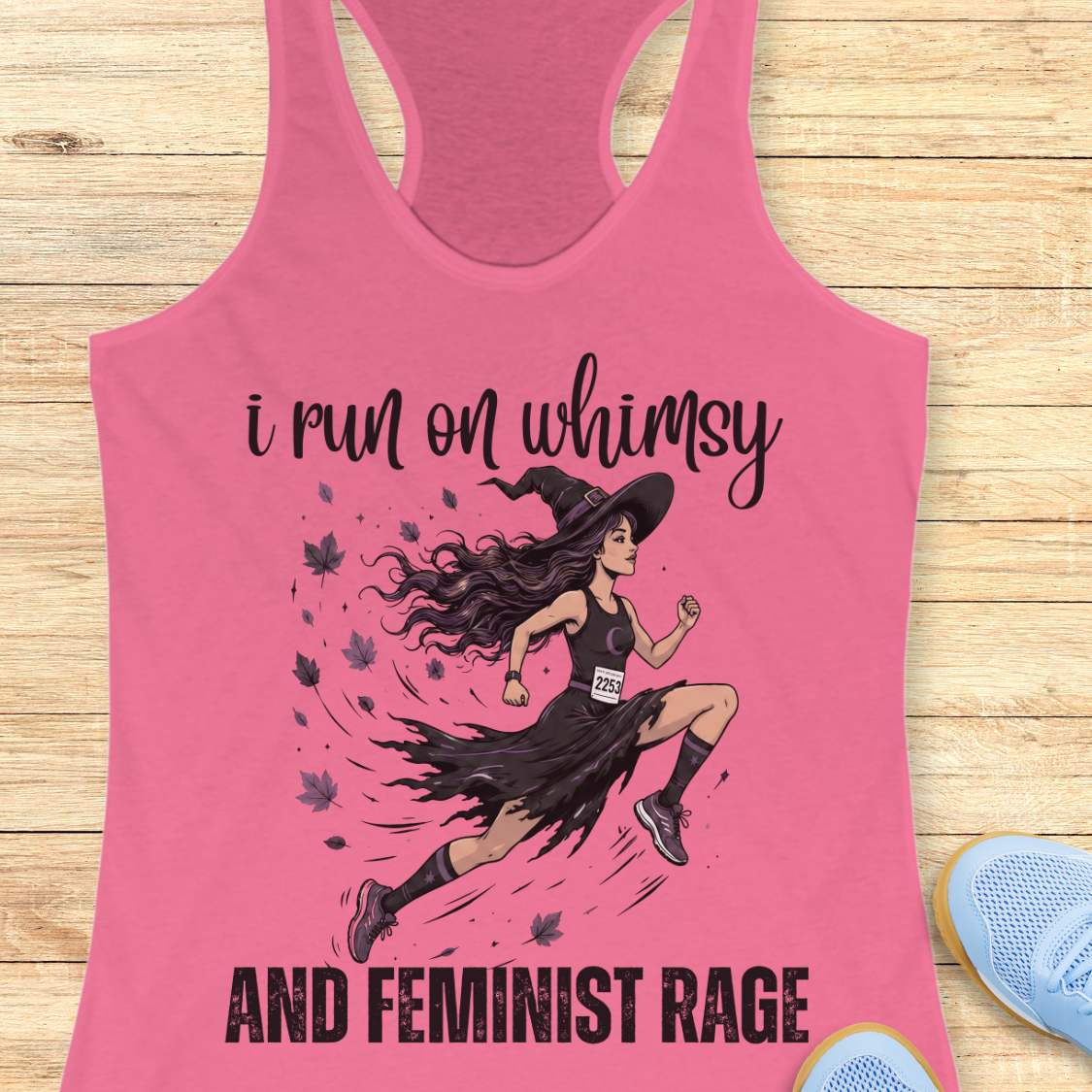 Pink tank top with a graphic of a woman running and text  i run on whimsy and feminist rage, on a wooden surface