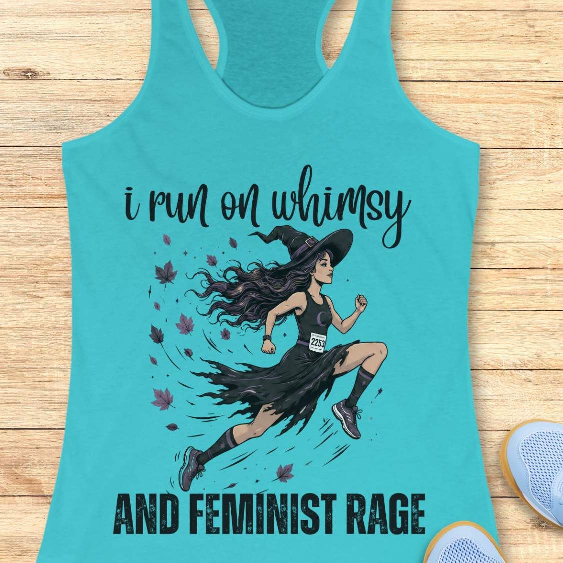 Turquoise tank top with a graphic of a witch running and text  i run on whimsy and feminist rage, on a wooden surface